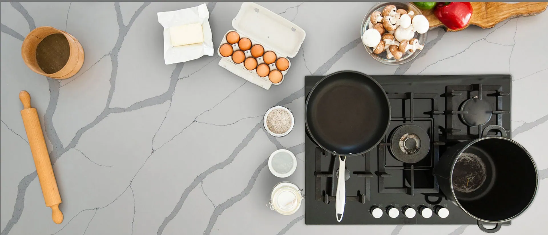 A top-down view of a kitchen countertop with cooking items. Includes a rolling pin, salt in a wooden container, butter, eggs in a carton, mushrooms, peppers, and a stove with a frying pan and pot.