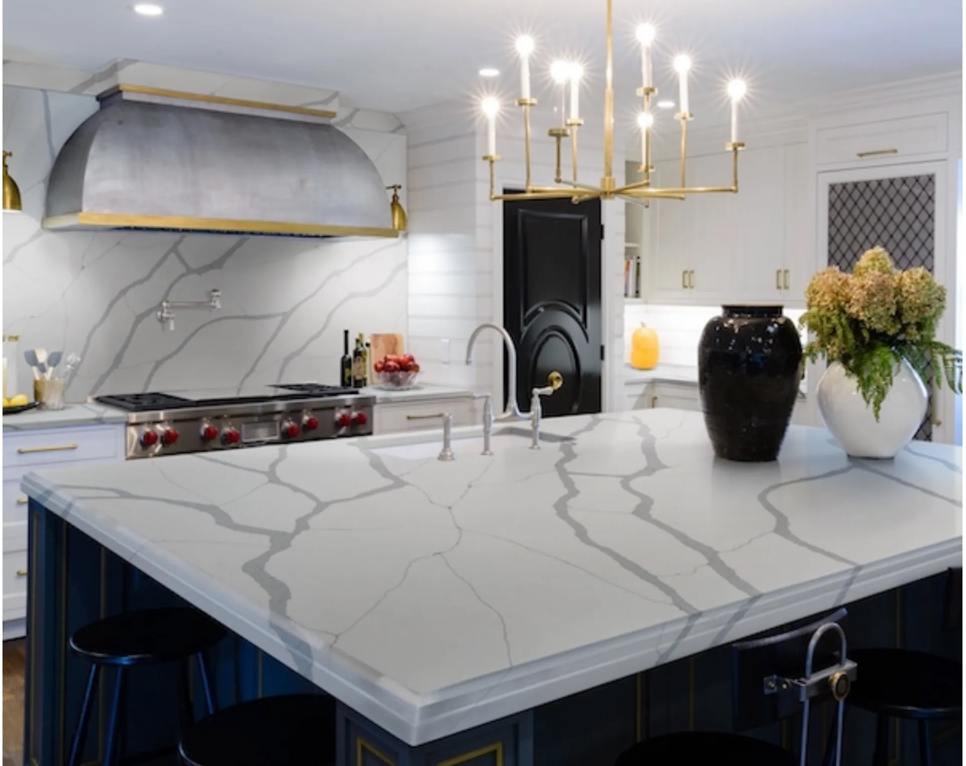 A modern kitchen featuring a large island with a white marble countertop and gray veining. A black vase with flowers sits on the island. The kitchen has white cabinetry, a stylish range hood, and a gold chandelier overhead.