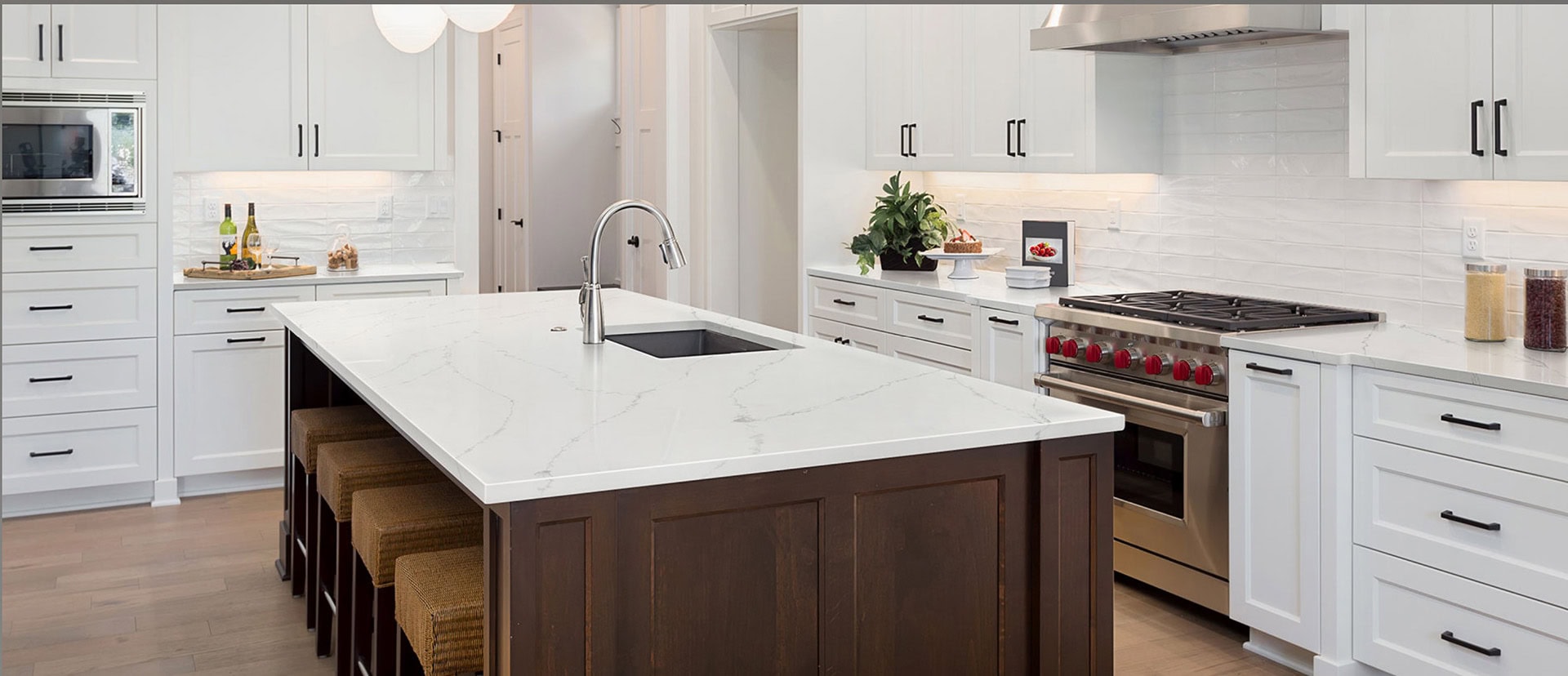Modern kitchen with a large white marble island featuring a dark wood base and four woven stools. Stainless steel appliances, white cabinetry, and a gas stove are present. Minimalist decor includes a fruit bowl and cooking essentials.