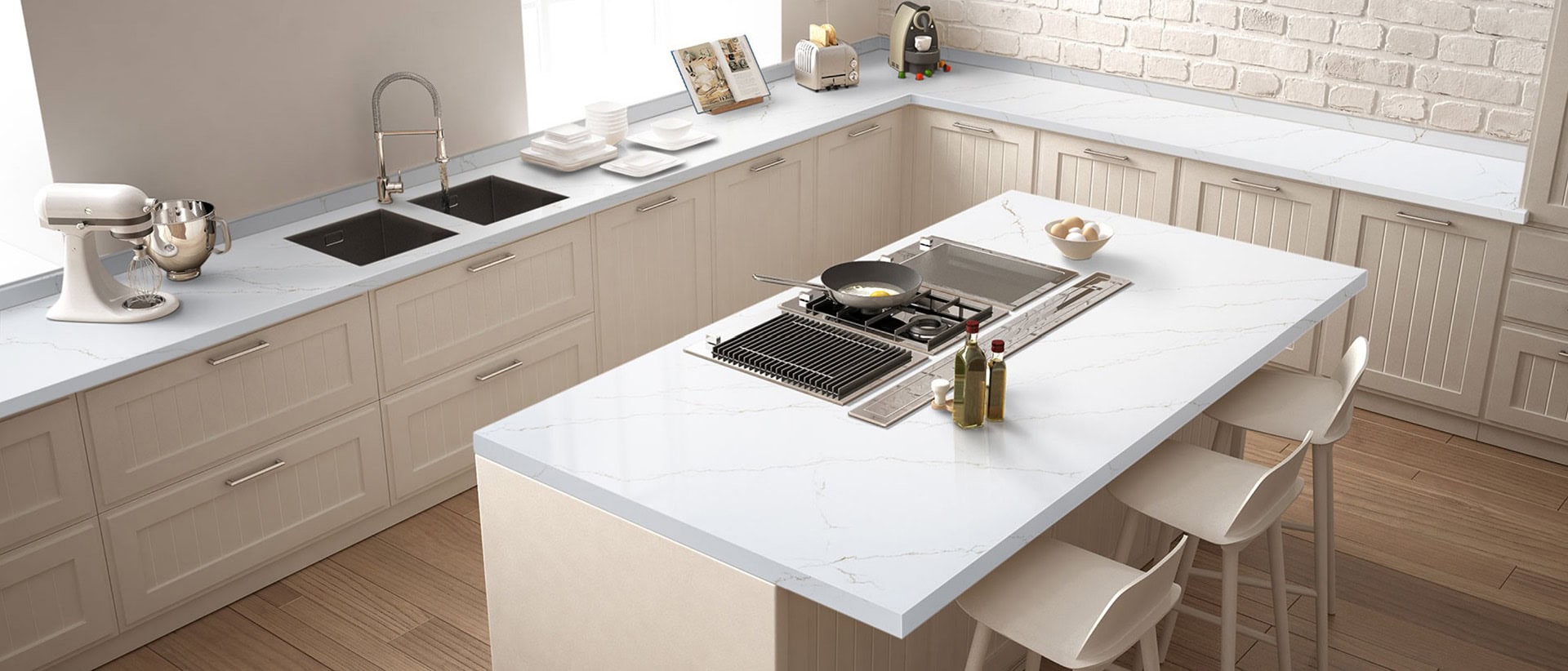 A modern kitchen with light wood cabinets and white marble countertops. A central island hosts a stove, pan, and condiments. A mixer, books, and dishes are on the counters. A window with soft lighting brightens the space.