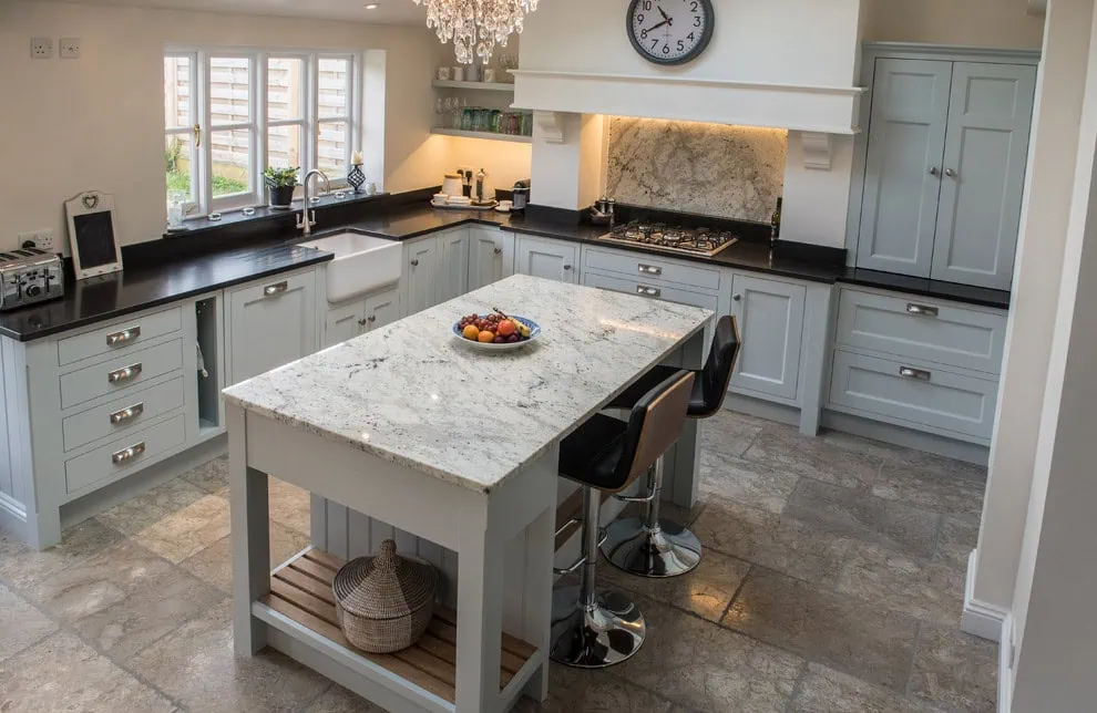 Modern kitchen with gray cabinets, marble countertops, and a central island with fruit bowl. Two bar stools are at the island. A chandelier hangs above, and a clock is on the wall. Natural light streams in through a window above the sink.