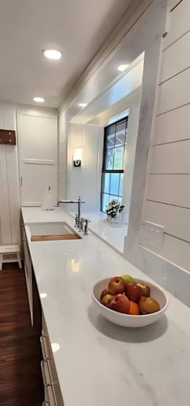 Modern kitchen with a white marble countertop, built-in sink, and stainless steel faucet. A bowl of apples sits on the counter. The walls are white with wood paneling, and a large window allows natural light to fill the space.