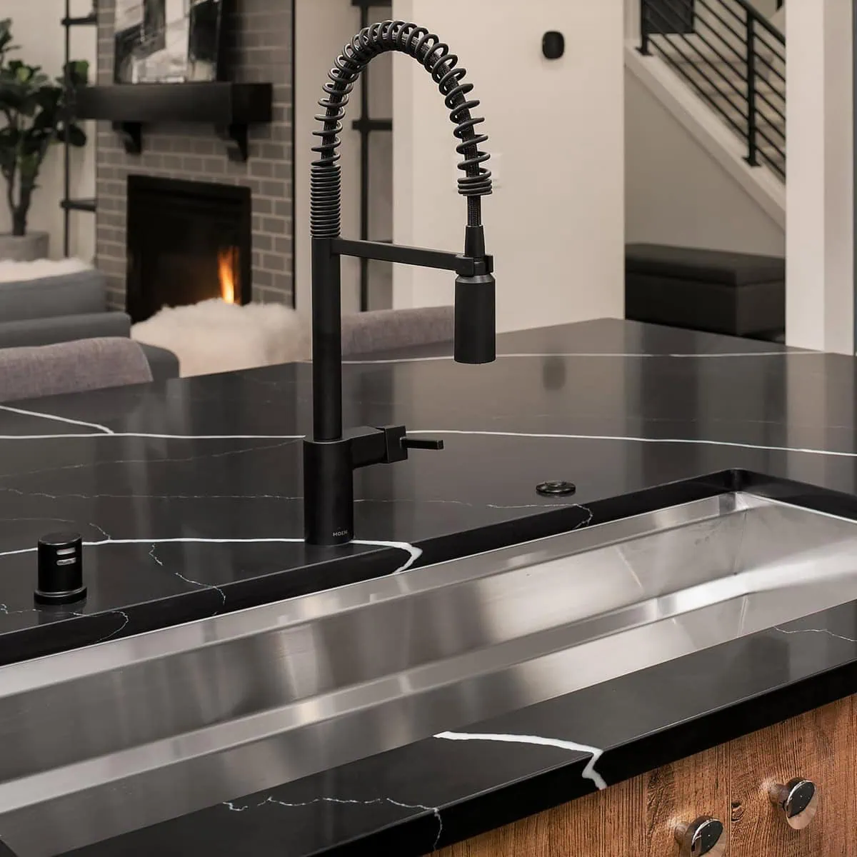 A modern kitchen with a black marble countertop featuring white veining. A sleek black faucet with a spring neck is mounted on a large stainless steel sink. In the background, there's a lit fireplace and a staircase with metal railings.
