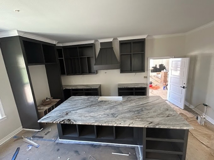 A modern kitchen under construction with dark cabinets, a marble island countertop, and an unfinished floor. A range hood is centered above the stove area. Light walls and an open door leading to another room can be seen. Tools and materials are scattered.