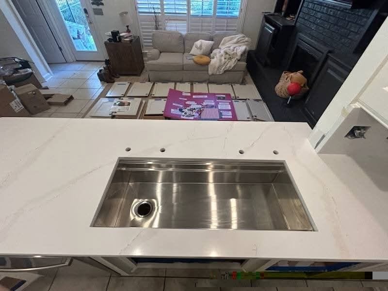 A white kitchen island with a stainless steel sink and multiple faucet holes. The background shows an open living area with a light-colored sofa, a fireplace, and a scattered pink board with papers. Unpacked boxes are visible on the tiled floor.
