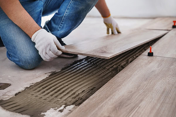 A person wearing jeans and white gloves is installing light brown wooden flooring. They are placing a plank over adhesive on the floor, surrounded by tools and spacers.