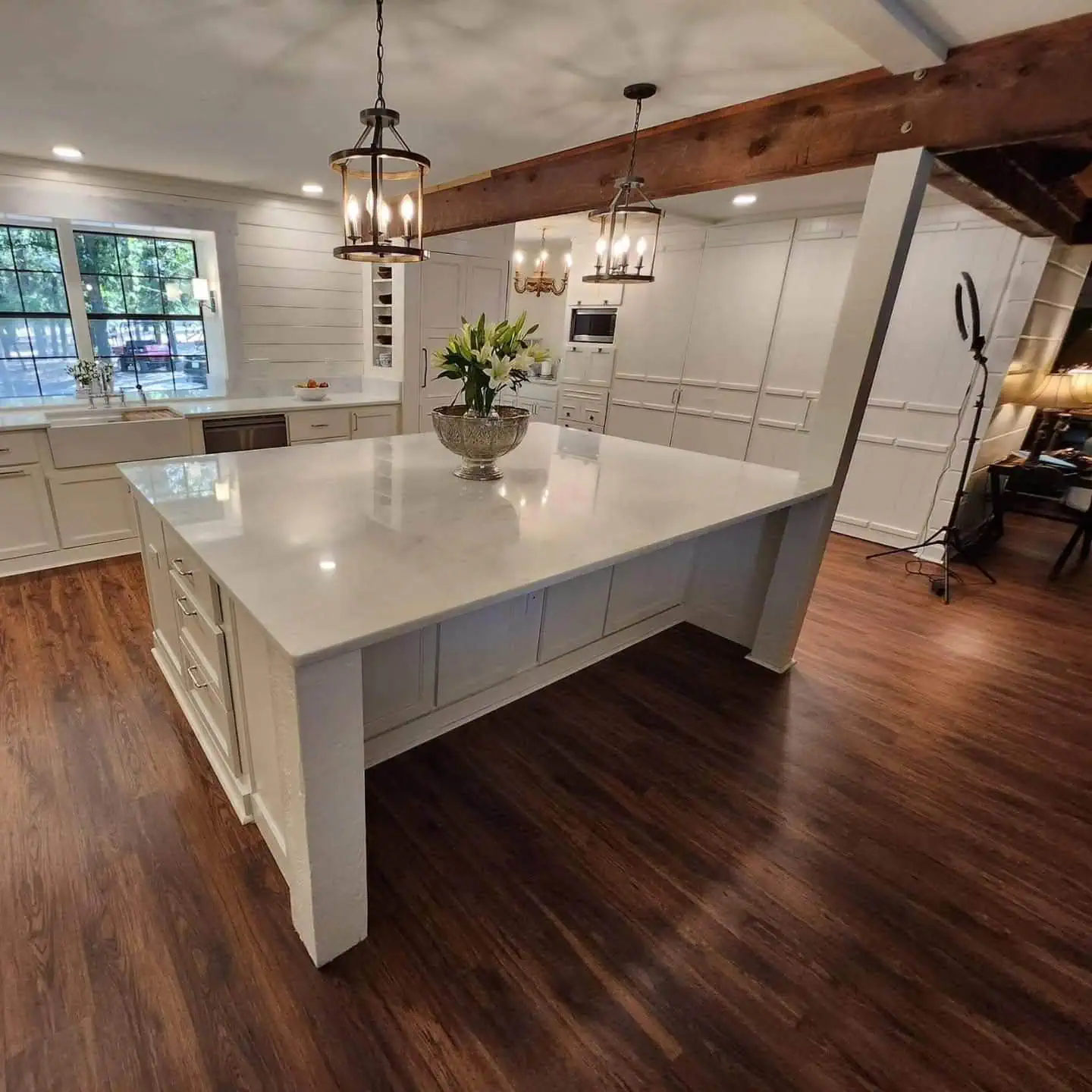 Spacious kitchen with a large white island and a vase of flowers. Dark wood floors and white cabinets add contrast. Two pendant lights hang above, and a window offers natural light. An exposed wooden beam accents the ceiling.
