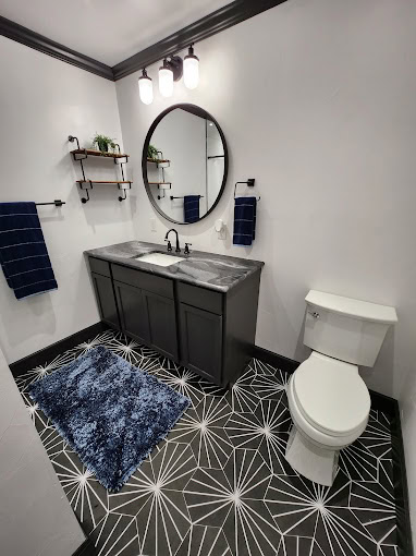 Modern bathroom with black vanity, round mirror, and three-bulb light fixture. White walls, black and white geometric floor tiles, and a white toilet. Blue rug, navy towels on racks, and two small plants on wall-mounted shelves.