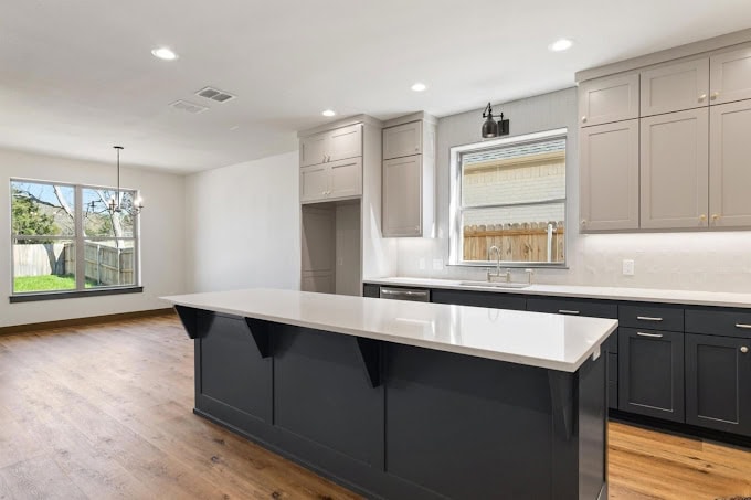 A modern kitchen featuring a large island with a white countertop and dark cabinetry. Light gray cabinets are mounted above, with a window and sink beneath. Wood flooring and recessed lighting add warmth and brightness to the space.