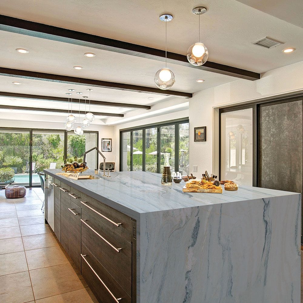 Modern kitchen with a large marble island featuring wooden drawers. The island is set with bread, pastries, and a small tray. Three pendant lights hang overhead. Sliding glass doors provide a view of a lush garden and pool area.