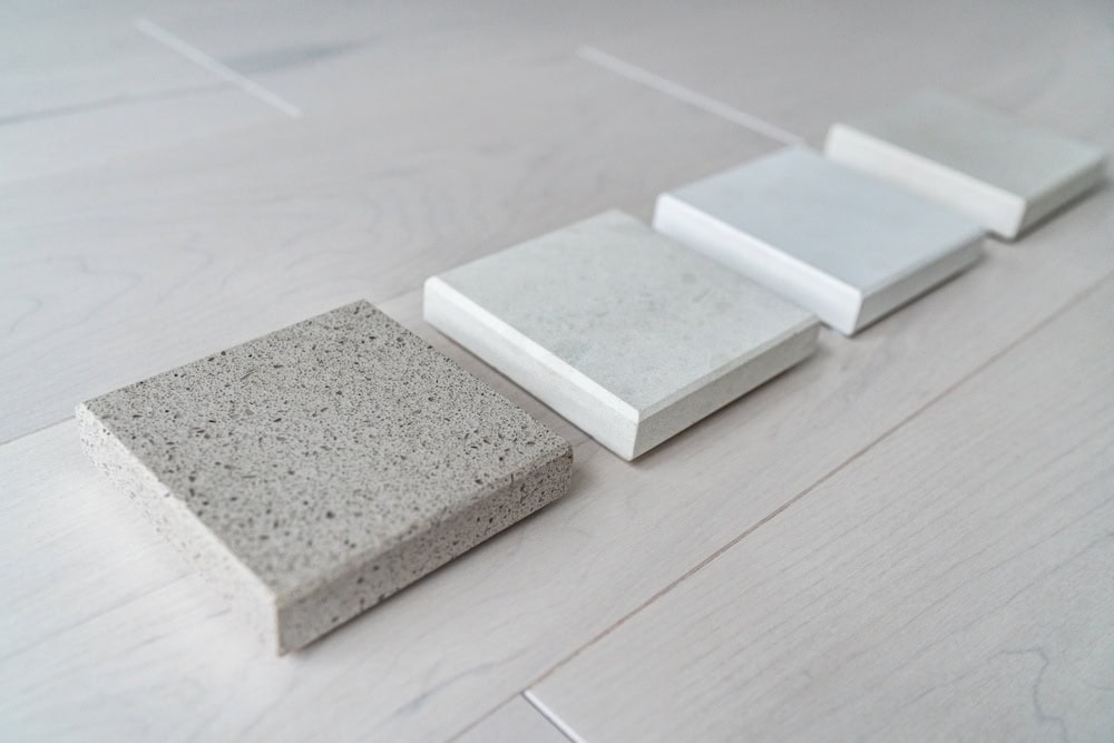 Five square stone samples in various shades of gray are arranged in a diagonal line on a light wood surface. The textures range from speckled to smooth, showcasing a variety of finishes.