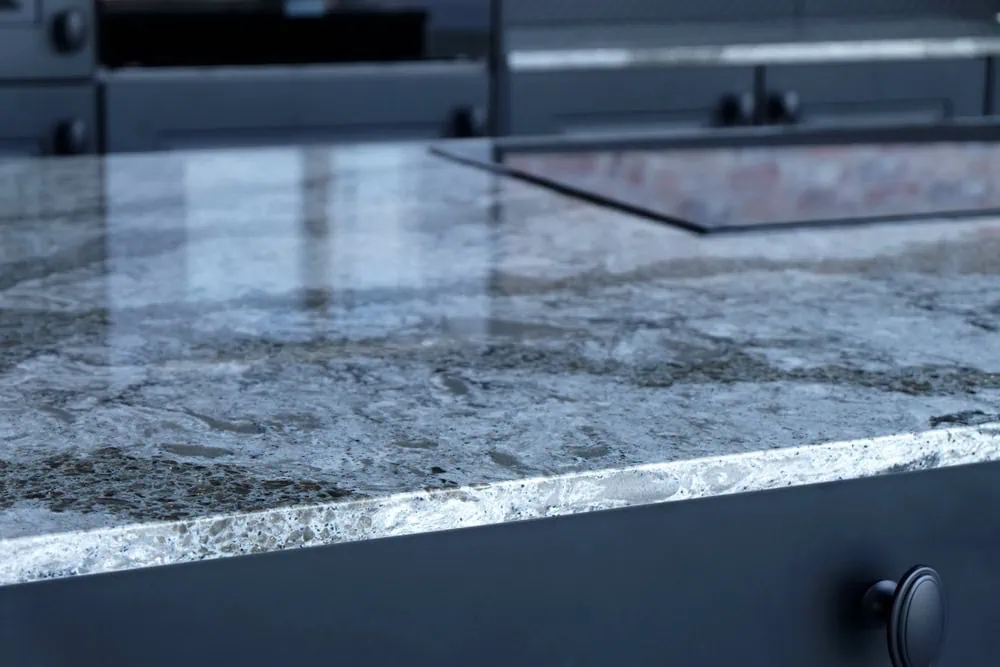 Close-up of a sleek, modern kitchen countertop with a shiny, gray stone surface. The edge features a polished finish, and a black induction cooktop is embedded on the right side. The background shows dark cabinetry.