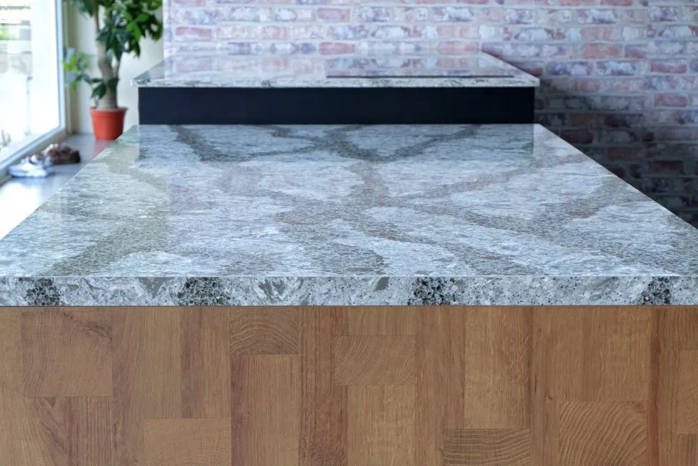 A modern kitchen island featuring a glossy gray granite countertop with unique veining patterns. The base is constructed from light-colored wood. A potted plant and brick wall are visible in the background, creating a cozy atmosphere.