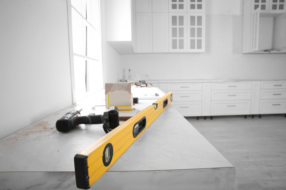 A home kitchen under renovation with white cabinetry and marble countertops. A yellow spirit level and a cordless drill rest on the countertop, alongside some construction materials.