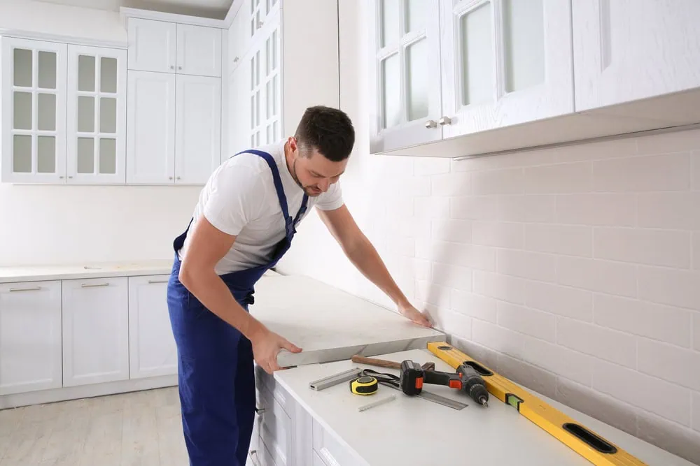 A person in blue overalls installs a countertop in a modern white kitchen. Various tools such as a level, measuring tape, and screwdriver are on the counter. White cabinets with glass panels are visible in the background.