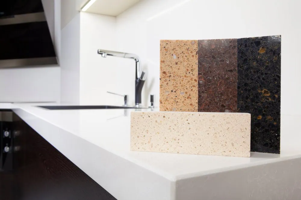 Kitchen countertop display with three stone samples in varied colors and patterns. There's a faucet and sink in the background on an L-shaped white counter, all within a modern kitchen setting.