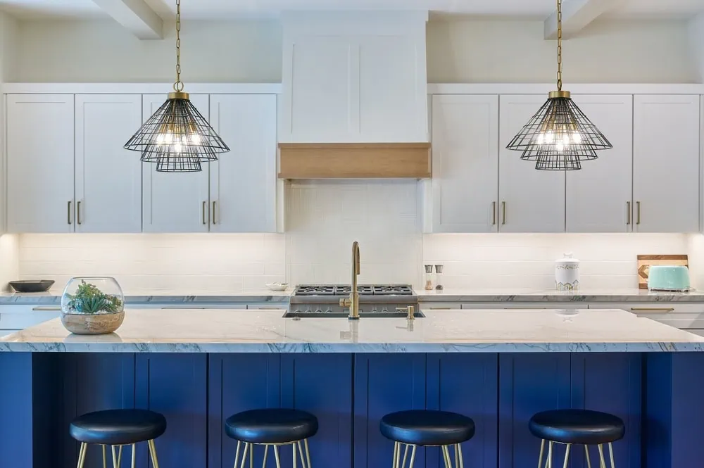 Modern kitchen with a large blue island featuring a marble countertop and four black stools. Two wire pendant lights hang above. White cabinets with gold handles line the back wall, and a wooden range hood is centered above the stove.