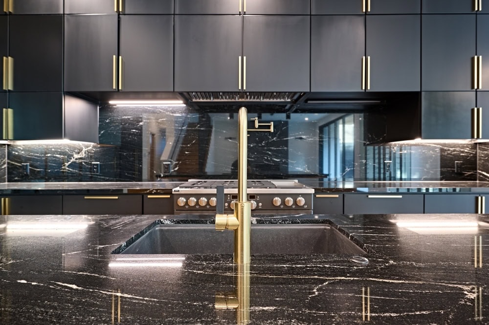 Modern kitchen with sleek black cabinets and brass handles. A black marble countertop features a built-in sink with a brass faucet. The backsplash mirrors the countertop. Recessed and strip lighting illuminate the space.