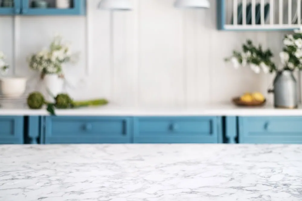 A kitchen with a marble countertop in the foreground. The background features blue cabinets, white flowers in vases, lemons in a bowl, and hanging pendant lights, creating a fresh and inviting atmosphere.