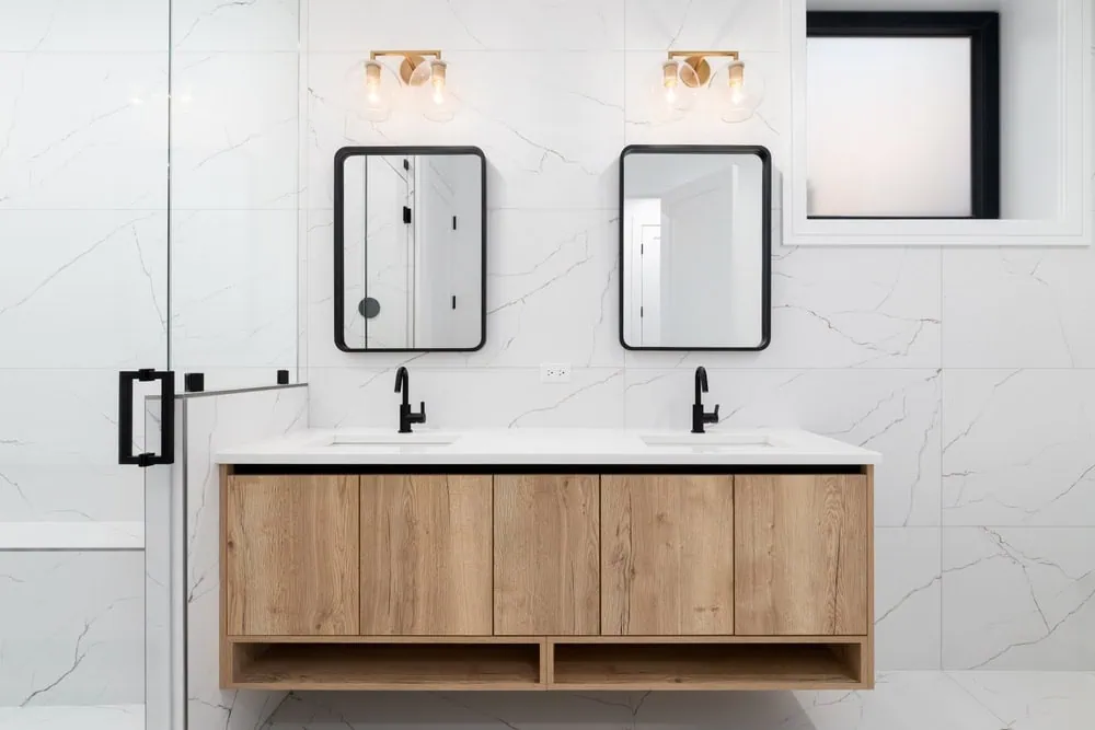Modern bathroom with a wooden double vanity, white countertop, and black fixtures. Two rectangular mirrors with black frames are above the sinks. The walls are white with subtle gray veining, and glass shower doors are visible on the left.