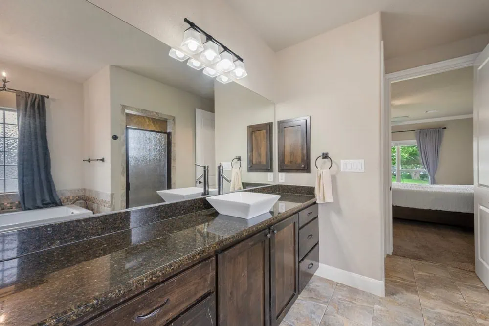 Modern bathroom with a dark granite double vanity, square white sinks, and a large mirror. A shower with a glass door and a tub are visible. Towels hang on the wall, and a doorway leads to a bedroom with a bed and gray curtains.