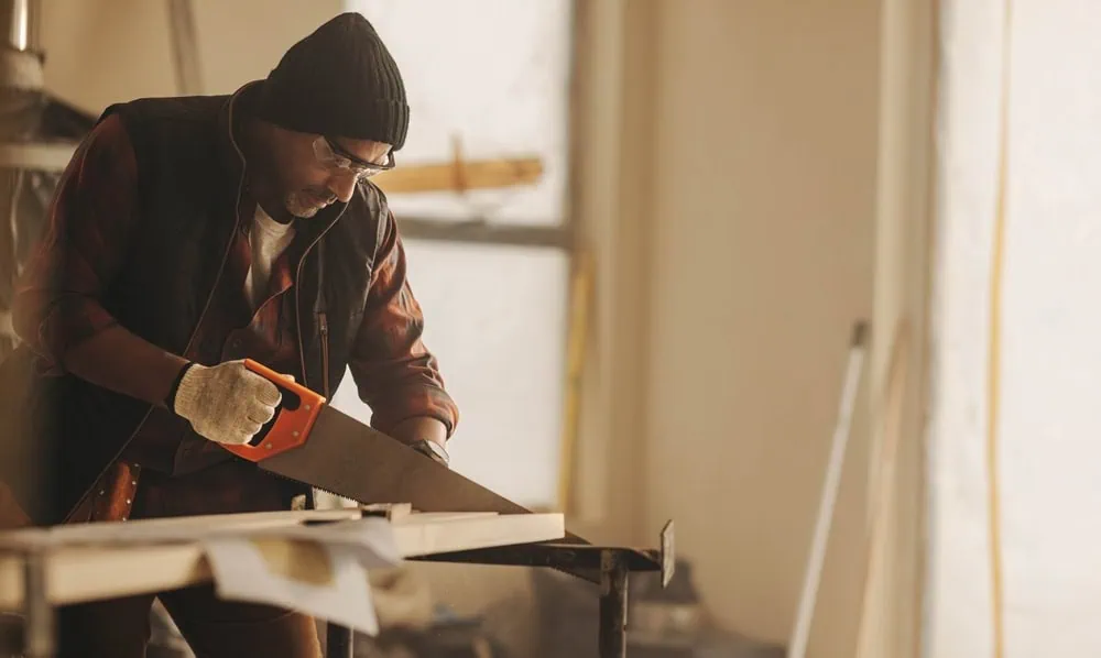 A man in a beanie and safety glasses uses a hand saw to cut a wooden plank on a workbench. He's wearing a plaid shirt, vest, and gloves, in a workshop with soft lighting. Various tools and materials are visible in the background.