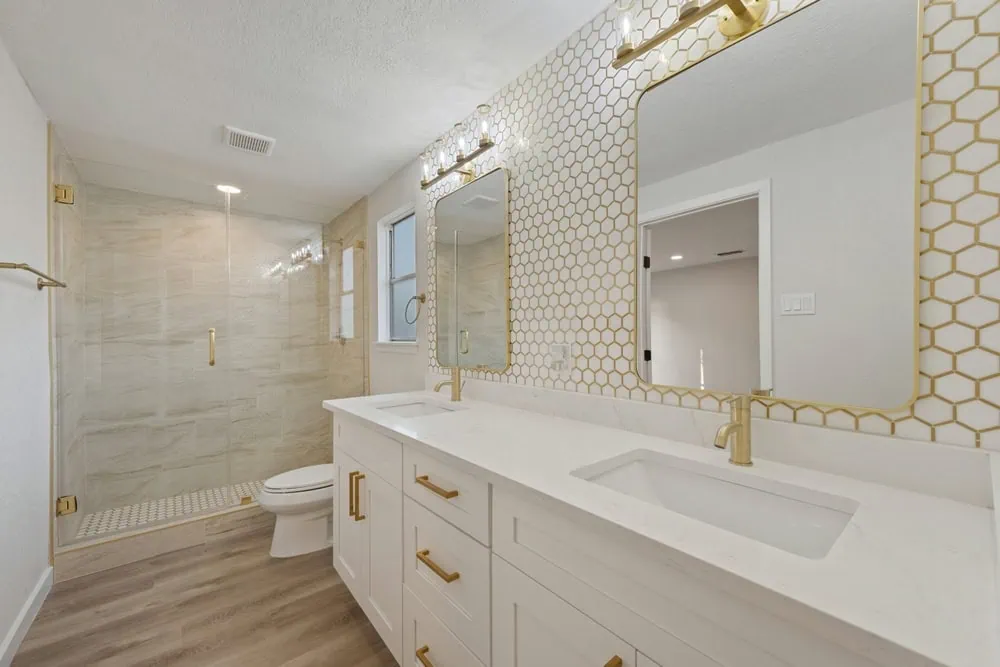 Modern bathroom with dual sinks on a white vanity, gold fixtures, and hexagonal tile backsplash. A large mirror hangs above the vanity. To the left, there's a glass-enclosed walk-in shower and a toilet. Light wood flooring adds warmth.