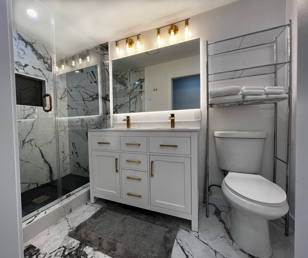 Modern bathroom with marble-patterned tiles, featuring a glass-enclosed shower. It has a white vanity with dual sinks and brass fixtures. Above is a lit mirror. On the side, a toilet is situated under a metal shelf stocked with towels.