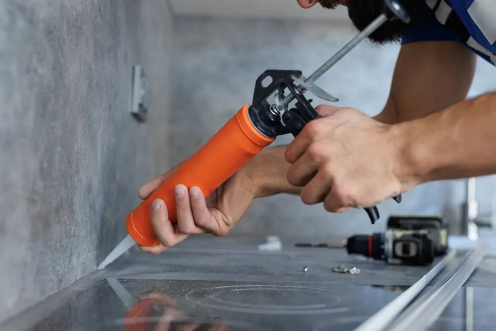 A person applies sealant with an orange caulking gun along the edge of a kitchen countertop. The surface is gray, and a drill is visible in the background. The scene suggests home improvement or maintenance work.