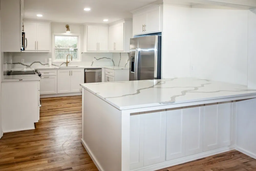 Modern kitchen with white cabinets and marble countertops. Stainless steel appliances, including a refrigerator and oven, are visible. The kitchen features wooden floors and recessed lighting, creating a bright and open atmosphere.