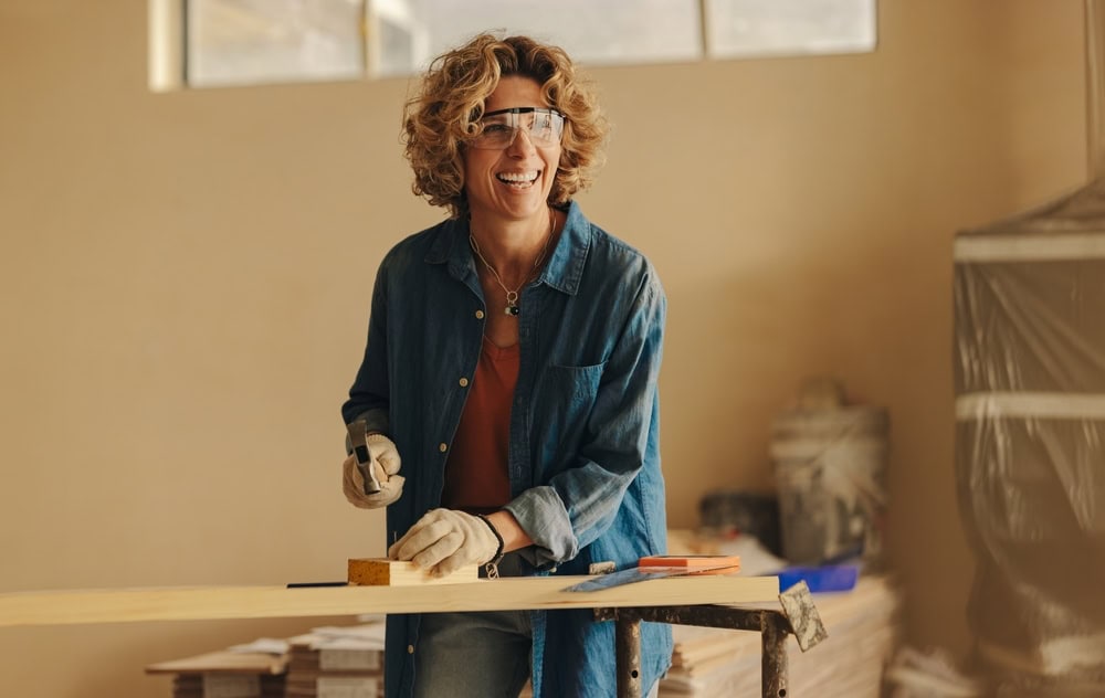 A person with curly hair wearing safety goggles and a denim shirt saws a wooden plank with a handsaw. They are standing at a workbench in a bright room and appear to be smiling. Tools and materials are visible on the bench.