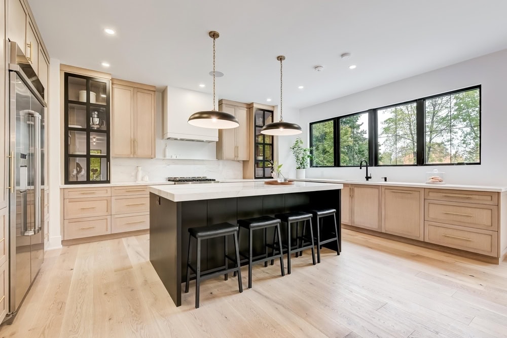 Spacious, modern kitchen with light wood cabinets, black island with three stools, large windows, white countertops, pendant lights, and stainless steel appliances. The room features light wood flooring and minimalistic decor.
