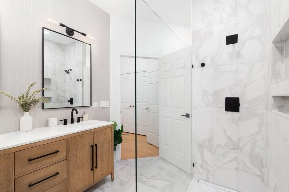 Modern bathroom with wooden vanity and large mirror. Features black fixtures, a white countertop, and a walk-in shower with marble tiles. A small plant decorates the vanity, and the room has a light, airy feel.