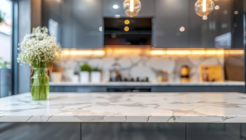 A modern kitchen with a marble countertop in the foreground, decorated with a vase of white flowers. The background features sleek, gray cabinets, a stovetop, and warm, under-cabinet lighting illuminating a marble backsplash.
