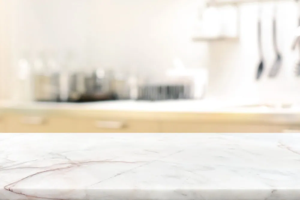 A marble kitchen countertop with a blurred background of kitchen utensils hanging from a rack and various glass jars on a countertop.