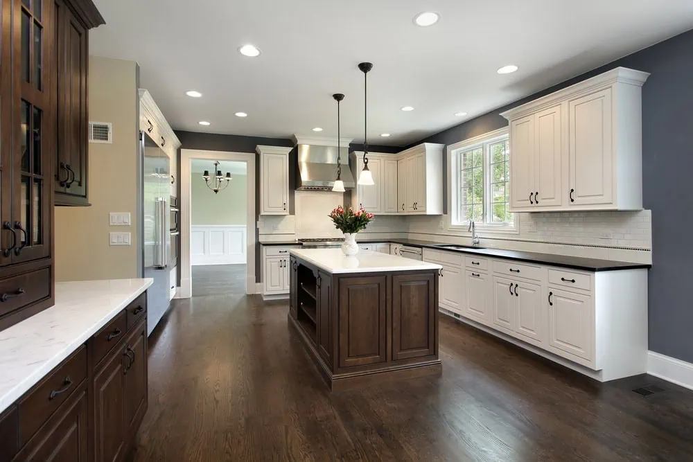 A modern kitchen with white cabinets, a dark wood island, and stainless steel appliances. Pendant lights hang above the island, which has a vase of flowers. Hardwood floors and a large window provide natural light.