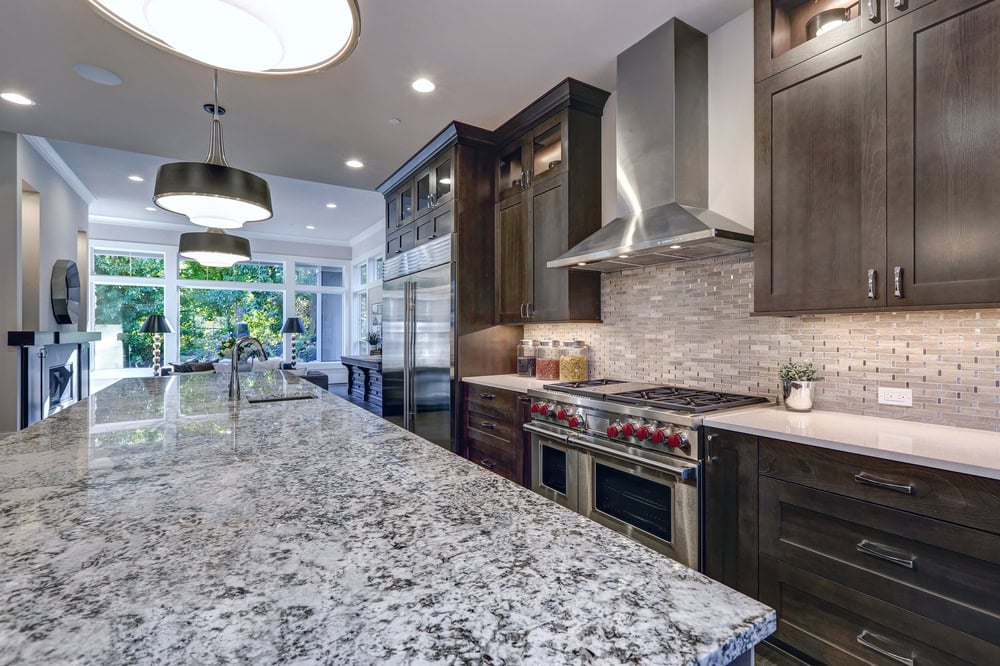 Modern kitchen with a large granite island, stainless steel appliances, and dark wood cabinets. A silver range hood is above the stove, and a tile backsplash adds detail. Bright windows provide natural light, with pendant lights over the island.