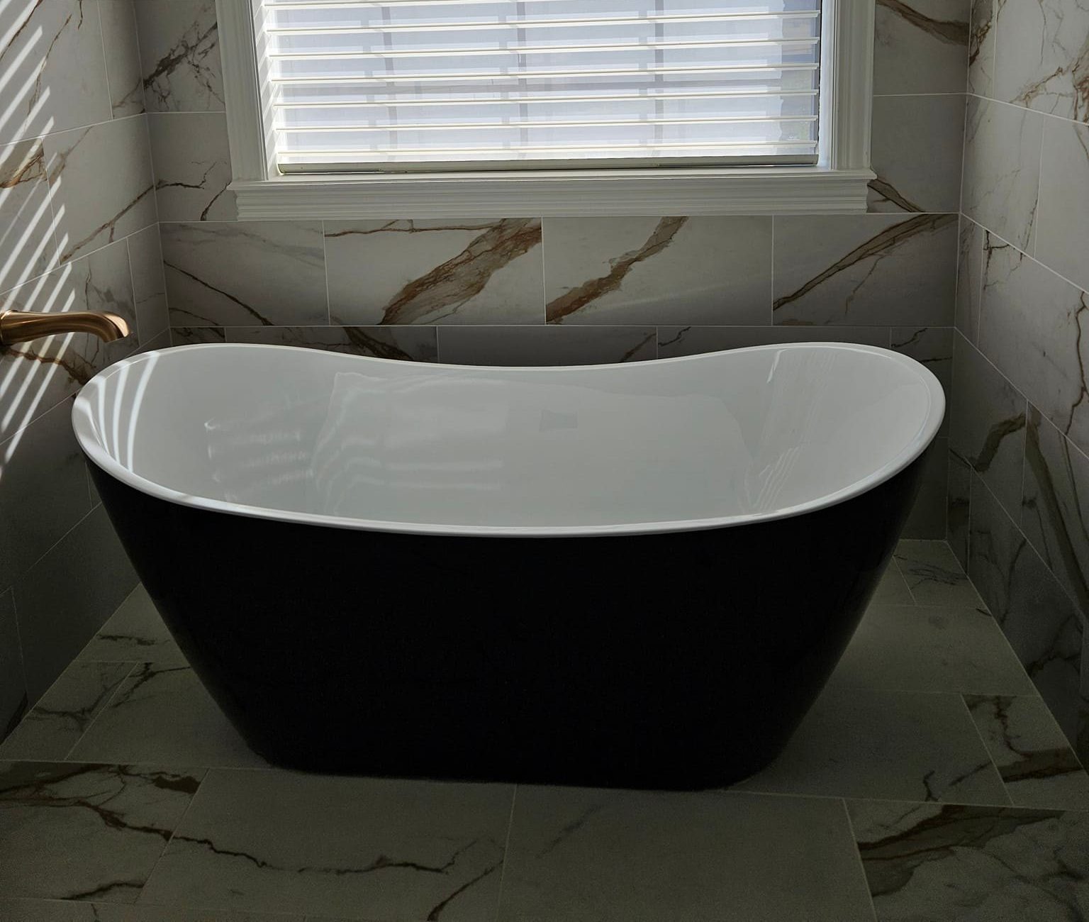 Freestanding black and white bathtub in a bathroom with marble-patterned tiles. A window with white blinds illuminates the room, casting light on the tub and surrounding area.