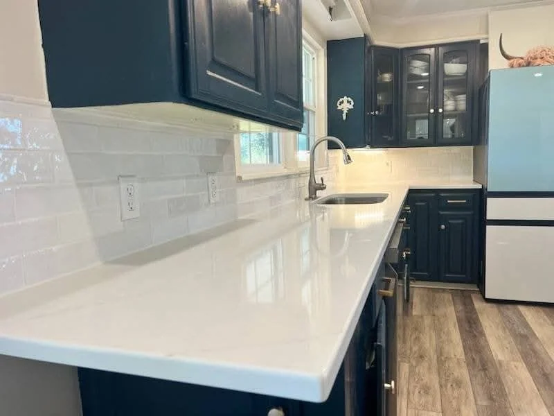 Modern kitchen with dark cabinets, white countertops, and a farmhouse sink. A stove is off to the side beneath a window, and natural light highlights the wood flooring. White tile backsplash adds contrast.