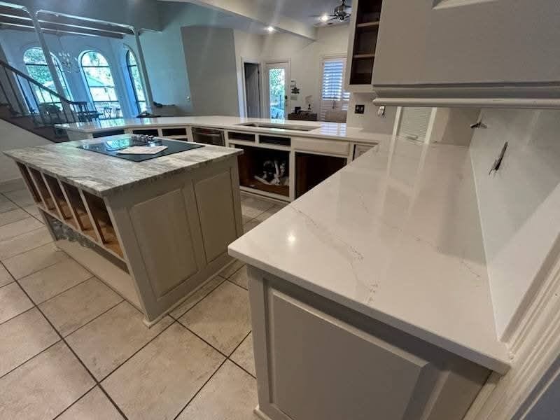 A modern kitchen features a marble-topped island with wooden storage underneath, set on a tiled floor. White countertops and cabinets are present, with arched windows and a staircase visible in the background.