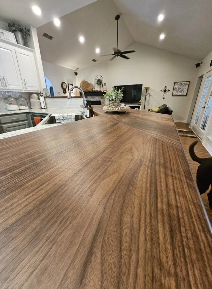 A spacious kitchen with a wooden countertop, stainless steel sink, white cabinetry, and recessed lighting. Spacious kitchen with wood countertop by countertop installers in Smith County, TX; cozy living area in the background.