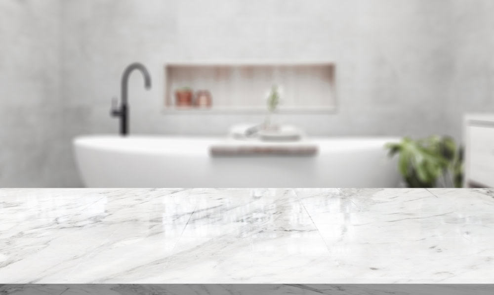 A modern bathroom features a marble countertop in the foreground. In the background, a white freestanding tub is partially visible, accompanied by a black faucet and small plants on a shelf.