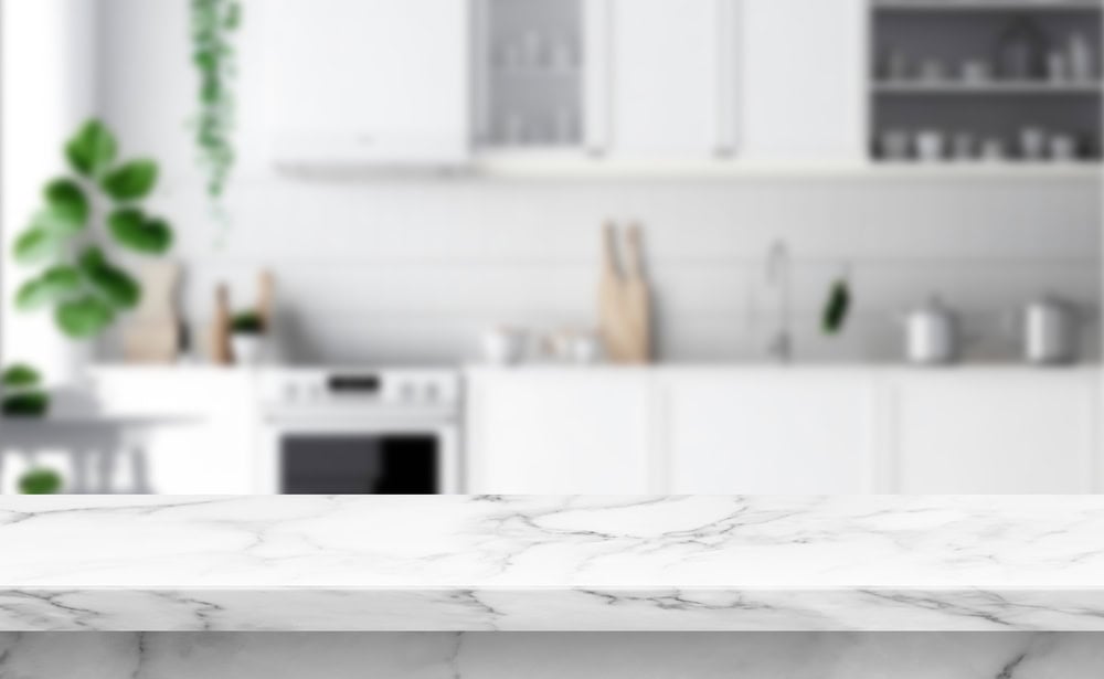 A modern, bright kitchen with white cabinets and appliances is softly blurred in the background. In the foreground, a clear marble countertop is in focus, adding elegance to the scene. A potted plant adds a touch of greenery.