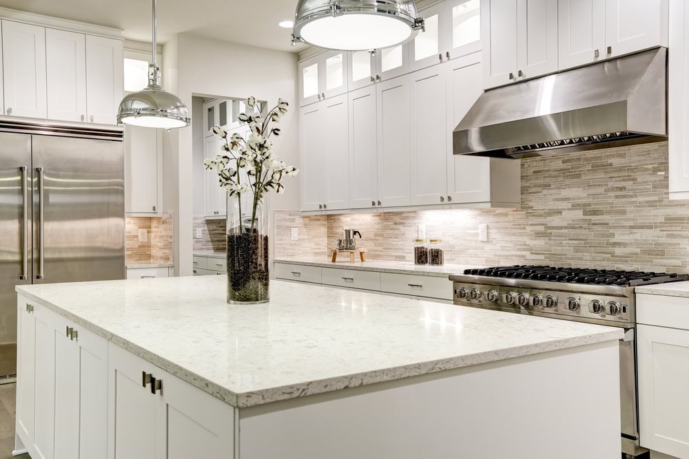 Modern kitchen with a large white marble island, stainless steel appliances, and pendant lights. The backsplash features a gray tile design. A vase with white flowers sits on the island, adding a touch of elegance.