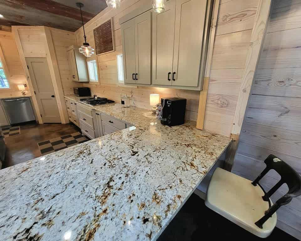 A kitchen with light wood walls, white cabinets, granite countertops, a stovetop, a coffee maker, and a white chair at the counter. Pendant lights hang from the ceiling, and sunlight enters through small windows onto expertly finished flooring installation.